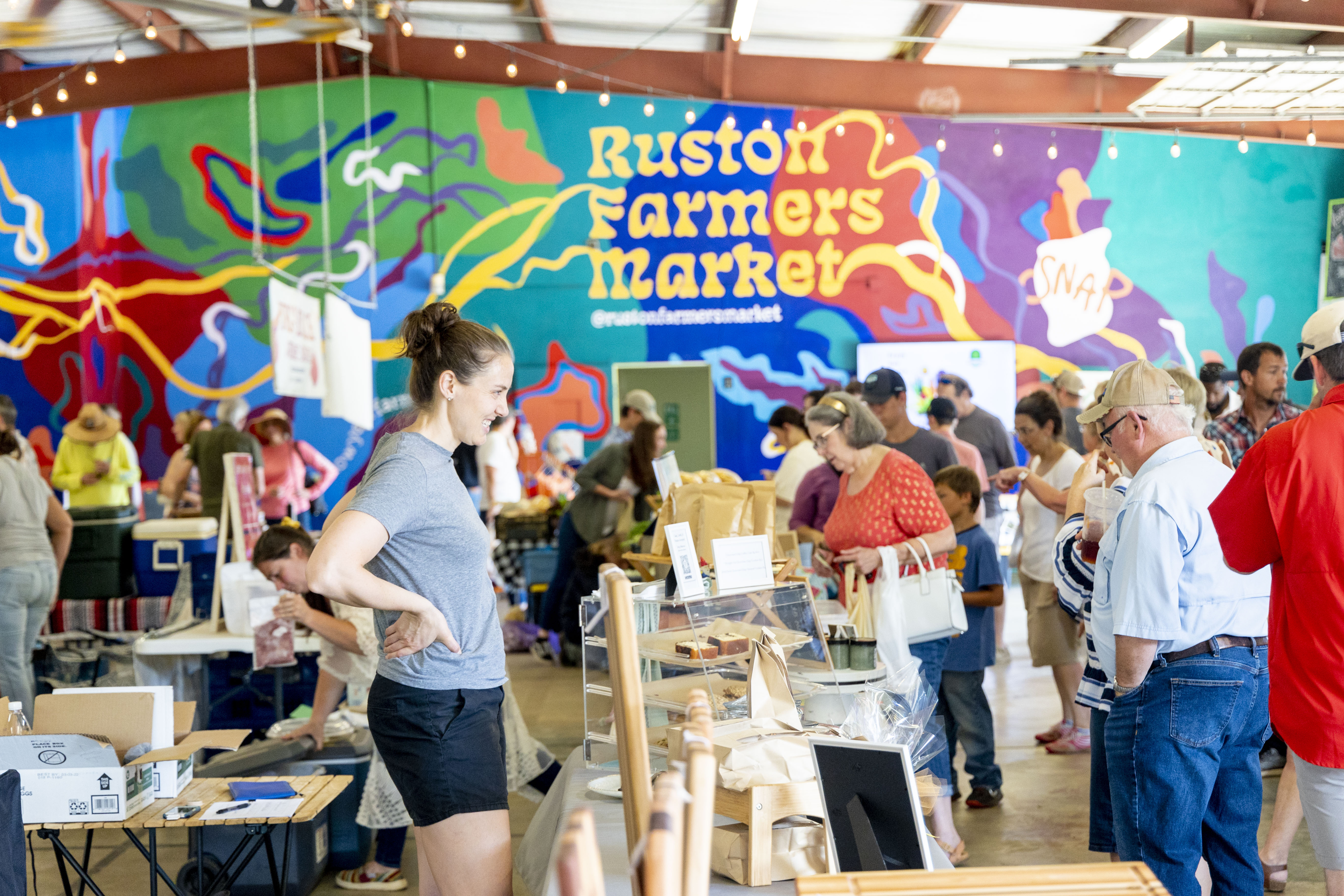Lively crowd shopping at the busy Ruston Farmers Market in Louisiana, with a colorful mural displaying “Ruston Farmers Market” in the background.