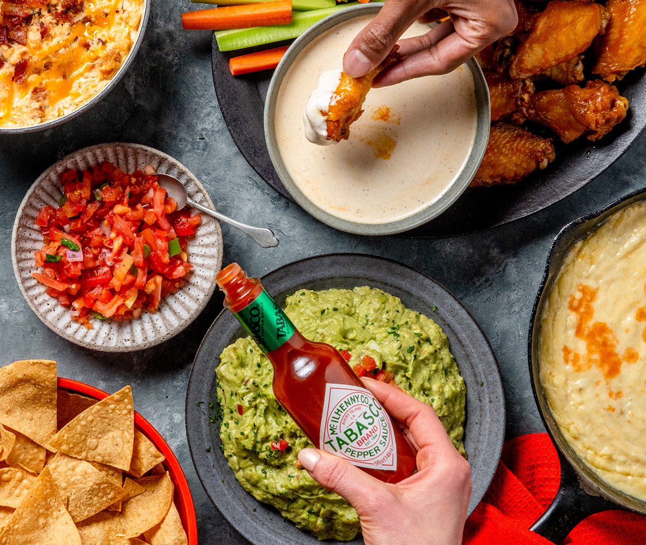 A Tabasco bottle is held over plates of sauces and other condiments, including guacamole and salsa.