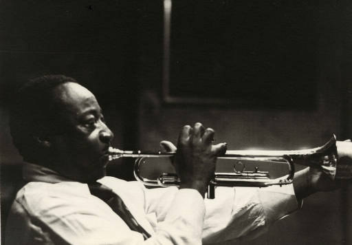 A side view of Dave Bartholomew blowing big cheeked into a trumpet in a dark room. 