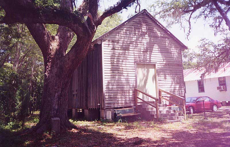 A white clapboard building sits beneath a canopy of trees. Stairs lead to a closed door on the front.