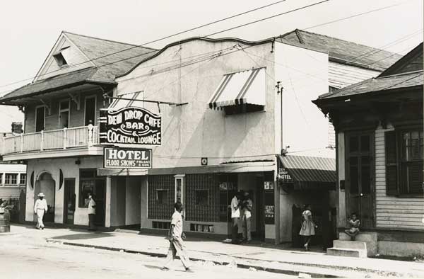 A black and white image of the exterior of the inn, with a sign reading "Dew Drop Inn"; a man crosses the street in front of it