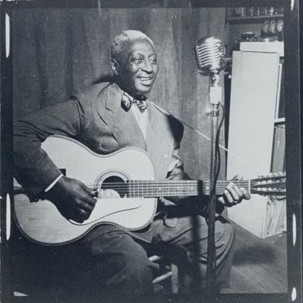 Leadbelly sits on a stool, strumming a guitar, while singing into a standing microphone