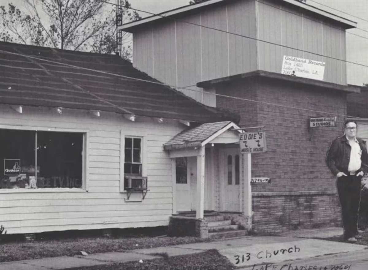 A man stands in front of two buildings, one a white clapboard house with a sign that says "Eddie's Music House," and one that's a double story, with a sign that says "Goldband Records."