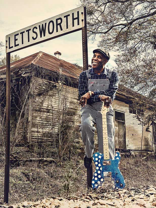 Buddy Guy poses with a blue guitar in front in front of a sign that says "Lettsworth"