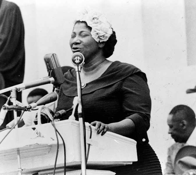 Mahalia Jackson sings from a podium on a stage. She wears a dark dress and has a fancy white hat on
