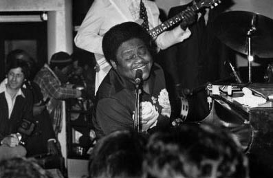 Fats sings into a microphone with a big smile on his face while playing the piano. A band plays behind him. 
