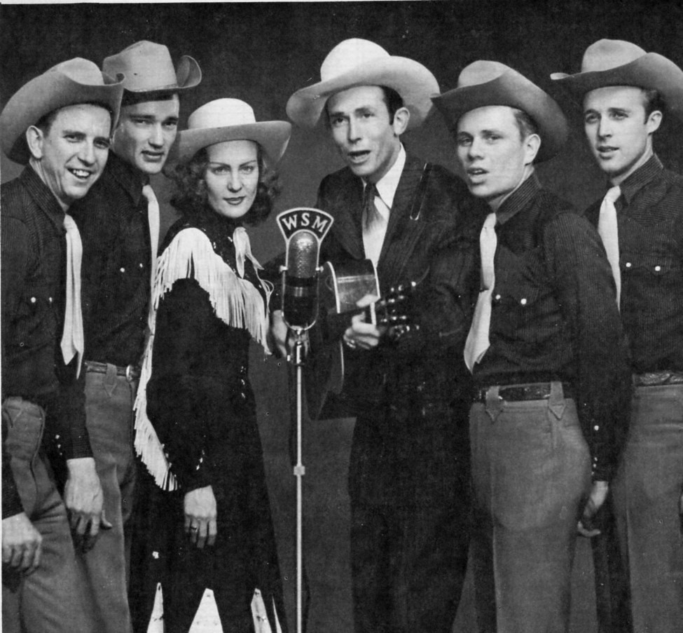 Five men and one woman in cowboy hats all sing into a microphone. They wear white ties and black shirts.