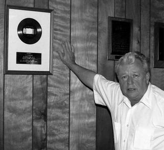J.D. Miller standing proudly next to the gold record of Kitty Wells's "It Wasn't God Who Made Honky Tonk Angels." Wikimedia. Photo by John Broven. 