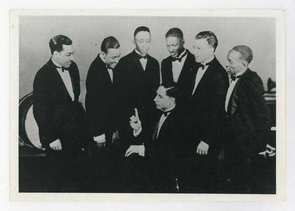 Jelly Roll sits with his finger in the air as if he's instructing his bandmates, all of whom stand in a circle around him. They all wear tuxedos.