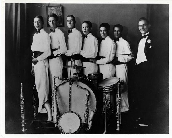 Seven men stand back to back behind a drum bit. The one on the far right wears a black tux; all the others wear white shirts and pants