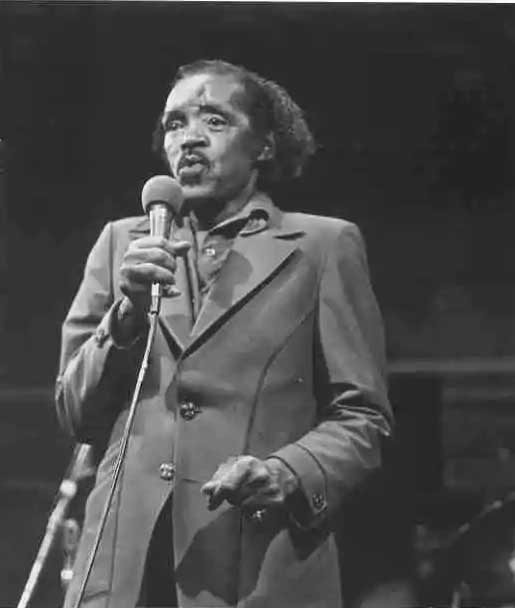 An older Percy wears and dark leisure suit and croons into a mic