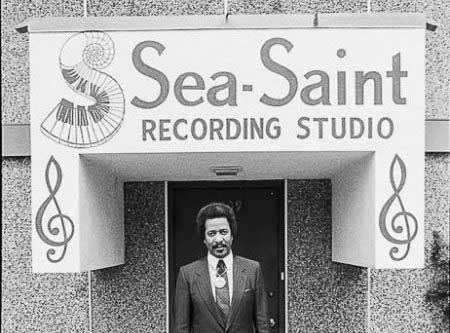Allen, in a black suit and tie, stands in front of the entrance to Sea-Saint Recording Studio. The sign for the studio features a swirling piano keyboard and two large music notes