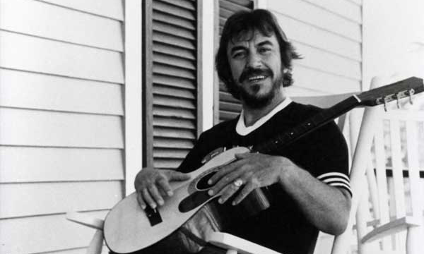 Bobby Charles sits on in a rocking chair holding a guitar on a home's front porch