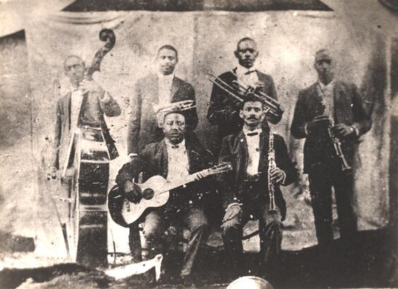 Buddy Bolder and his bandmates; four men stand with their instruments, and two sit; we see a base, guitars, trombones and clarinets