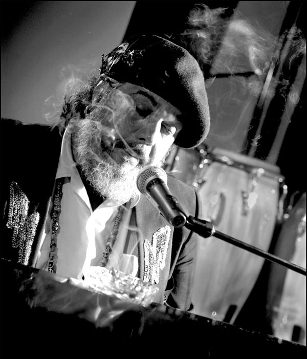 A tilted shot of Dr. John at a piano and mic. Smoke wafts around his head.