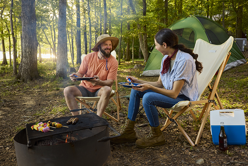 A couple sitting fire side at a camp site with tent in the background enjoying dinner at sunset