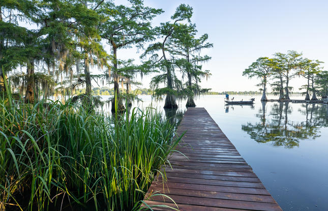 A wooden dock stretches into the water of Lake Bruin State Park, with a person fishing from a small boat in the background.