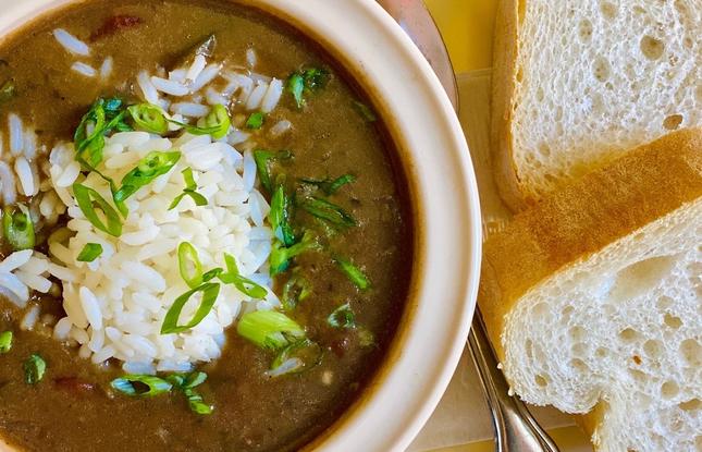 A cream colored bowl holds gumbo topped with a small mound of rice and sliced green onion. To the right of a bowl are to slices of French bread.