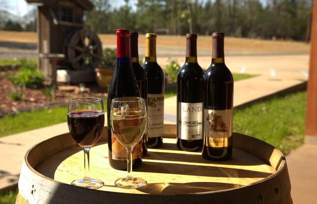 Bottles of red wine sit on a barrel behind a full glass of red wine and a full glass of white wine.