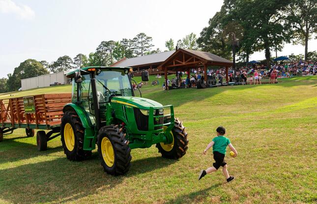 A child runs next to a tractor while families enjoy an outdoor concert in the background.