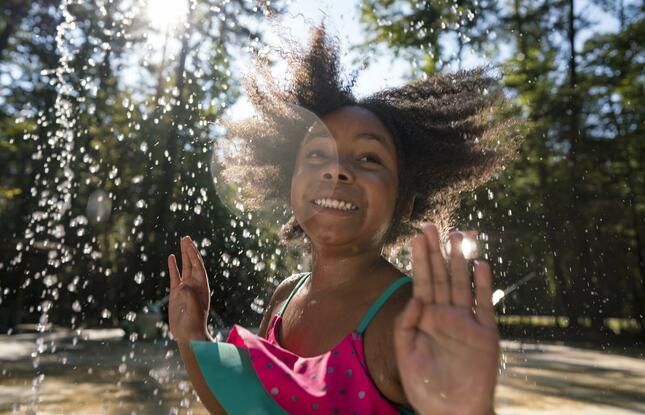A young girl smiling while she plays in a splash pad, with water spraying around her.