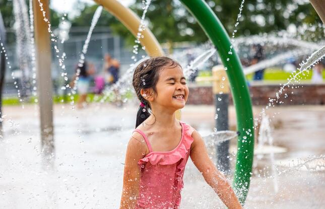 A little girl in a ruffled pink bathing suit smiles as she splashes around in a water playground at Highland Road Community Park in Baton Rouge, Louisiana.