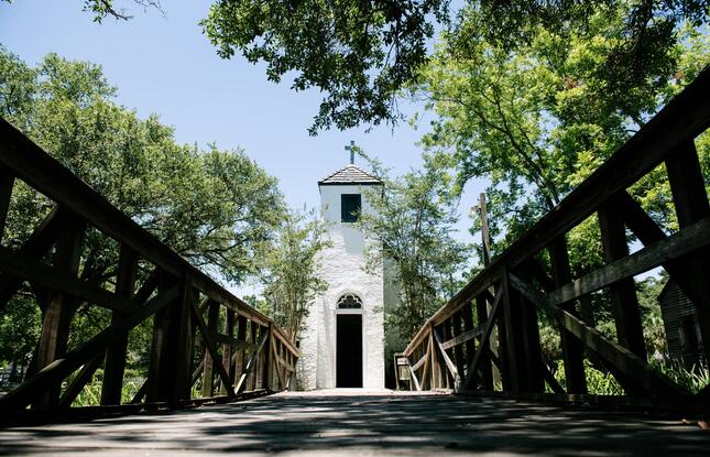 The white brick church at LARC’s Acadian Village, shot from the wooden bridge leading to the entrance on a sunny day.