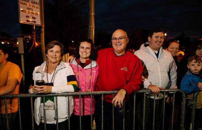 Four people wearing jackets and Mardi Gras beads smile for the camera while waiting for floats to pass by during a parade.