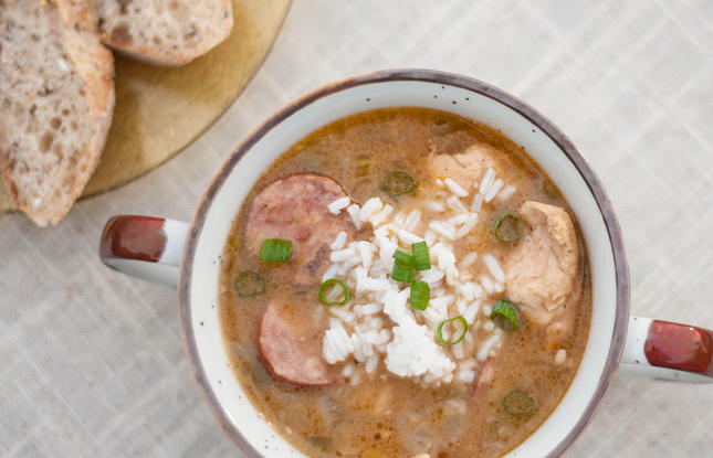 A white and brown mug of chicken and sausage gumbo, topped with white rice and sliced green onions, with slices of sausage and chunks of chicken visible through the well-seasoned broth.