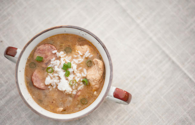 A white, red-rimmed mug filled with turkey and sausage gumbo, with chunks of white meat, slices of sausage, perfectly cooked white rice and sliced green onions.
