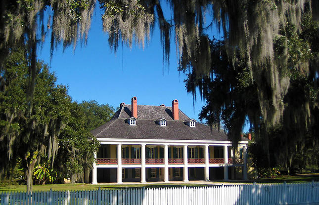 Front view of the Destrehan Plantation main house with white columns and moss-draped trees, surrounded by a white wooden fence.
