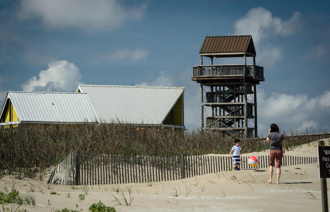 A mother and child play with a beach ball at Grand Isle Beach, with a lookout tower and sand dunes in the background.