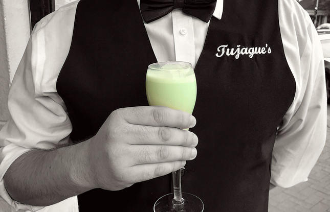 A server at Tujague’s holds a pale green Grasshopper cocktail in a stemmed glass, wearing a black vest and bow tie.
