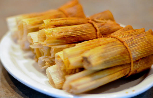 A white plate stacked with hot tamales, corn and meat wrapped in cornhusks.