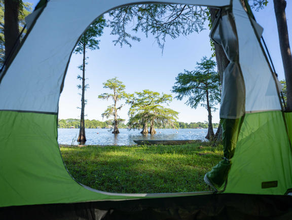 The flap of a green and blue tent opens up to reveal a beautiful lake with trees jutting out of the water at Lake Bruin State Park.