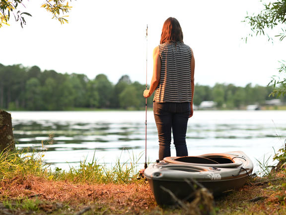 A person in a striped shirt and black pants stands on the shore holding a fishing pole next to a kayak at Jimmie Davis State Park.