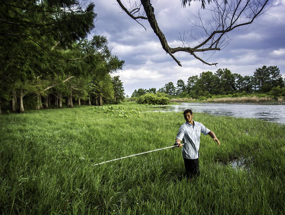 Man holding a fly-fishing pole and standing in marsh grasses on the banks of a river in Toledo Bend State Park in Louisiana.