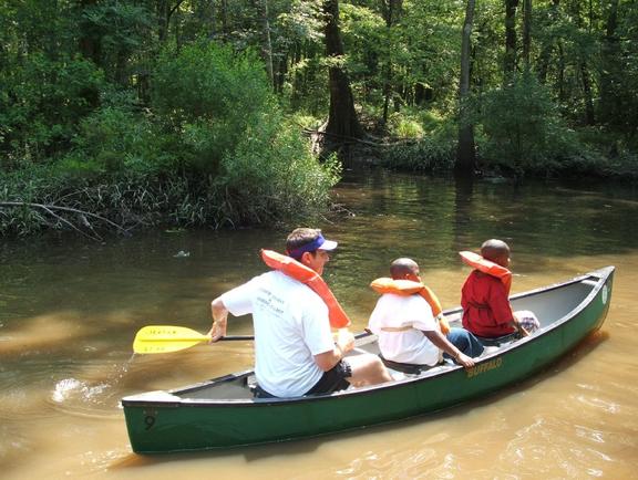 A man and two children wearing lifejackets paddle a canoe through the muddy waters of Tickfaw State Park.