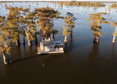 An aerial view of a wooden houseboat on the Atchafalaya Basin, anchored in a grove of massive trees.