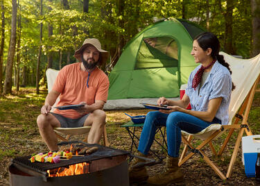 Couple cooking skewers over a campfire beside a green tent in a wooded Louisiana campsite.