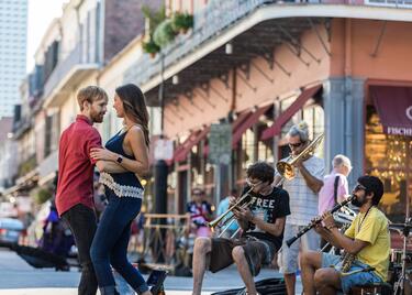 A couple dances in the street while a small brass band plays on Royal Street in New Orleans, with musicians seated and pedestrians watching in the French Quarter.