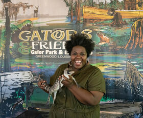 A woman in a green shirt holds a baby alligator in front of a sign advertising Gators &amp; Friends Adventure Park.