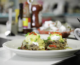 Eggs benedict, topped with shredded roast beef and green onions, served on a white plate, with condiments like ketchup and Louisiana hot sauce in the background, from Liz’s Where Y’at Diner.
