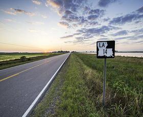 A rural two-lane highway, part of Louisiana's Cajun Corridor Byway, stretches into the distance. In the background the sun peeks over the horizon, brightening a cloudy sky.