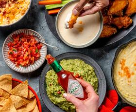 A Tabasco bottle is held over plates of sauces and other condiments, including guacamole and salsa.