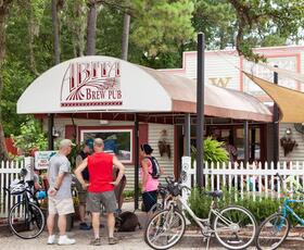Three people stand underneath an awning with a logo that reads, "Abita Brew Pub." Their bicycles are leaned against a white picket fence to the right of the awning.