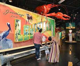 A woman looking at a large museum exhibit shaped like a postcard, with text that reads “Greetings from Louisiana” and has a red crawfish on top