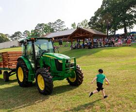 A child runs next to a tractor while families enjoy an outdoor concert in the background.