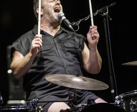 A drummer in a black collared shirt raises his drumsticks while singing at the Red River Revel Arts Festival.