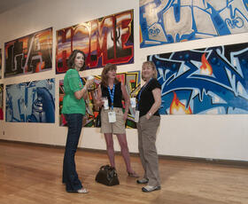 Three women stand and chat in front of several graffiti-style paintings at Artspace in Shreveport.